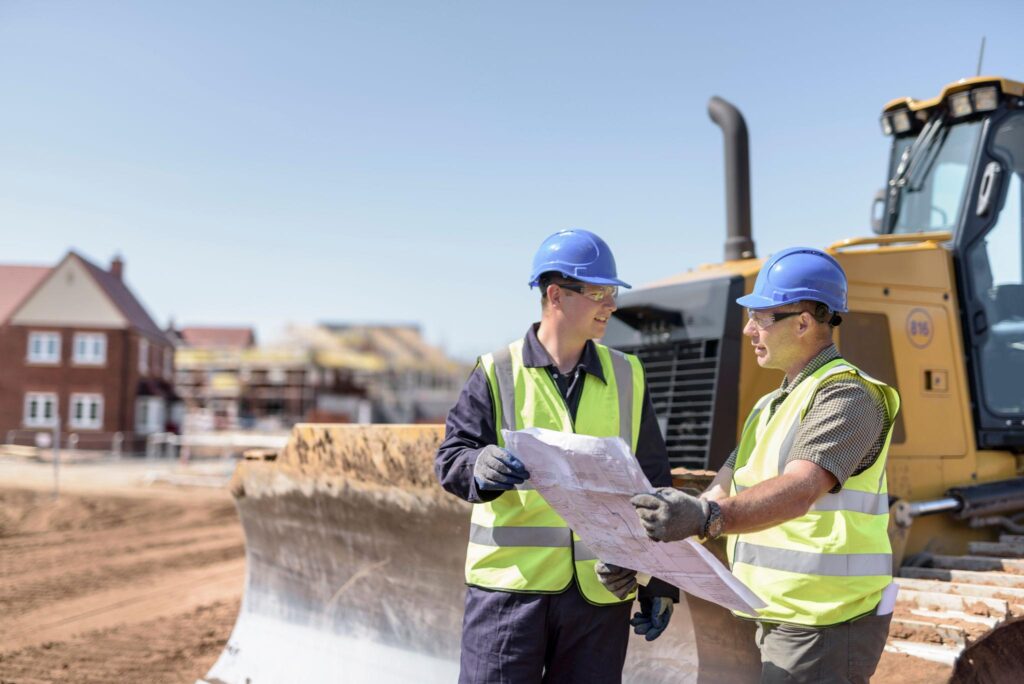 Builders reviewing construction plans beside survey equipment and heavy machinery during house staking preparation on a residential lot